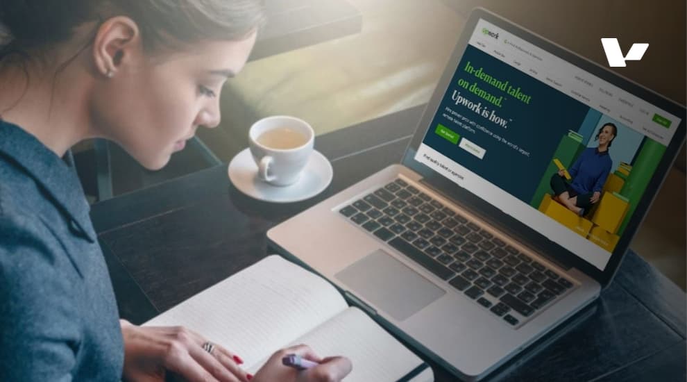 A person sitting at a desk, writing in a notebook while using a laptop with the Upwork website open on the screen.